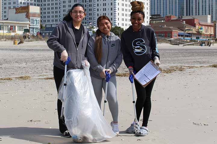 Three students with trash bags cleaning up a beach in AC