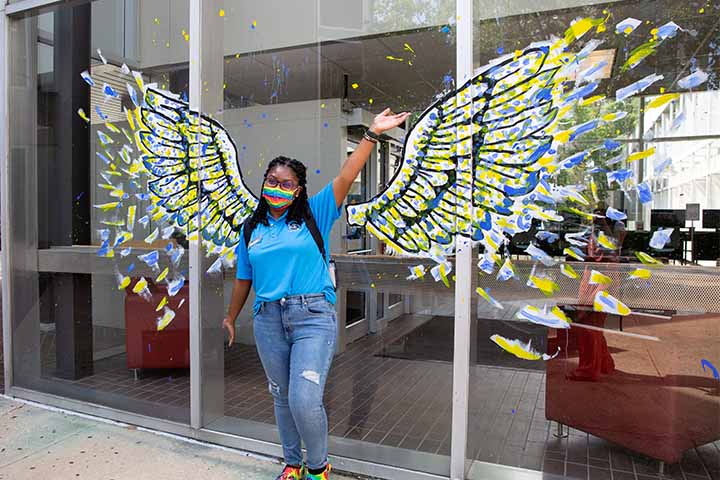 A student poses between blue and gold wings painted on a window