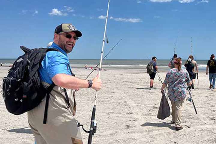 A man with a hat and fishing pole smiles while walking on the beach