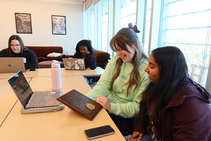 Four Stockton students sit at a table with laptops looking at the Nobel prize plaque 