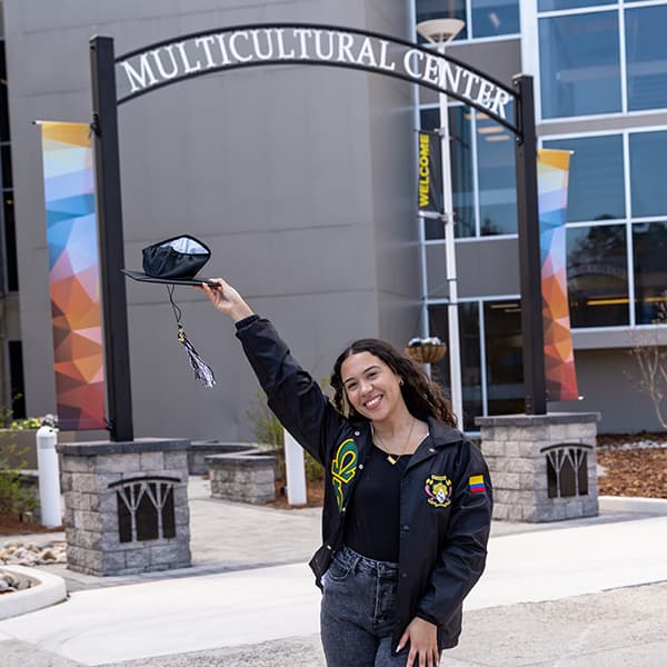 Britney Marrugo in front of the Multicultural Center arch, holding her graduation cap over her head