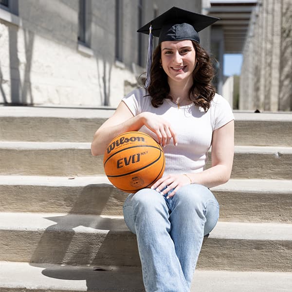 Emma Morrone sitting on the steps of the Campus Center, wearing her graduation cap and holding a basketball 