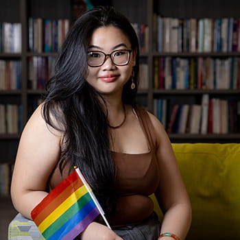 Van Nhi Ho smiling and sitting in the Women's Gender & Sexuality Center lounge with a pride flag in her hand
