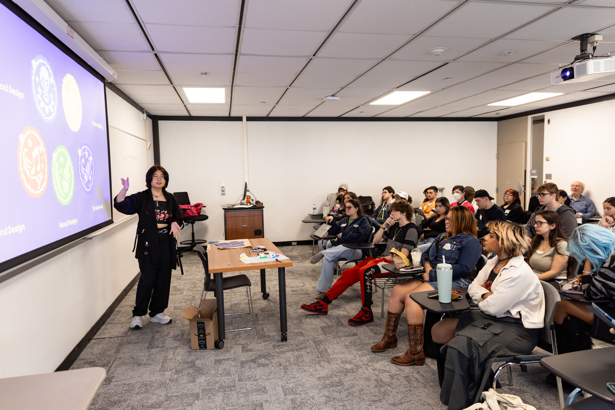 A person standing behind a table teaching in a classroom. The person is pointing to a slide presentation while a group of about 20 students listen attentively.