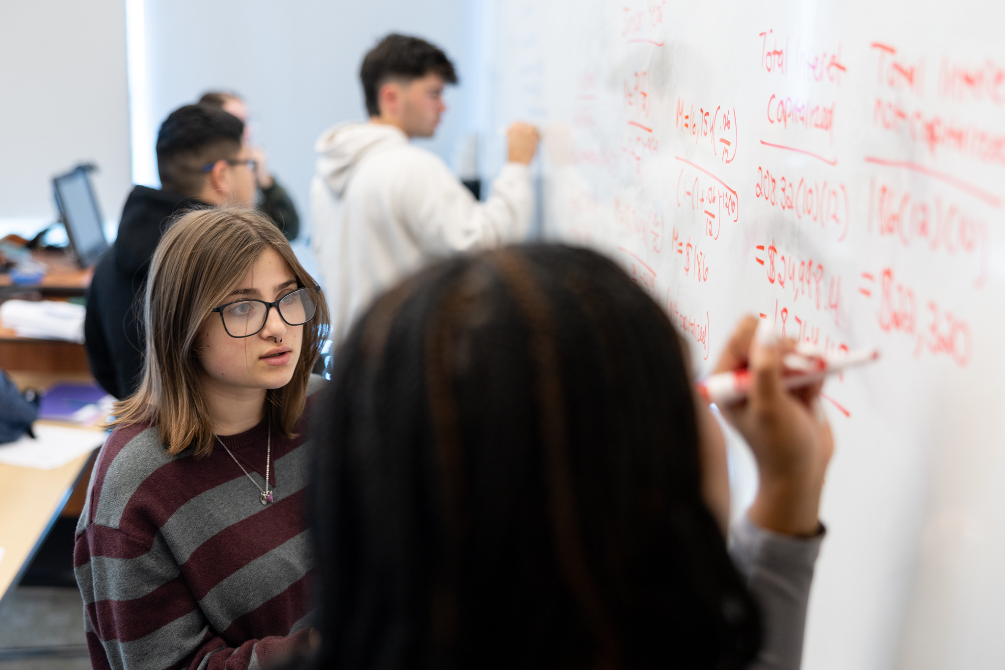A person wearing glasses working with another person to solve math equations on a white board. Three other people work on math equations in the background.