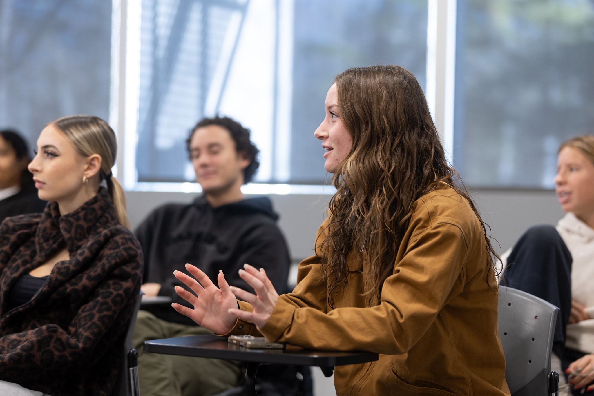 A person with long brown hair sitting in a desk explaining something. Three other people listen in the background.