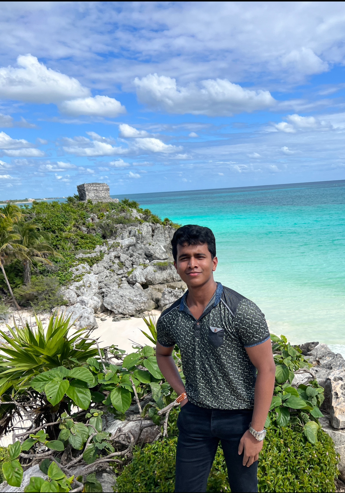Young man standing posing at camera with bright blue ocean water and a green grass-tipped beachy cliff in the backrgound.