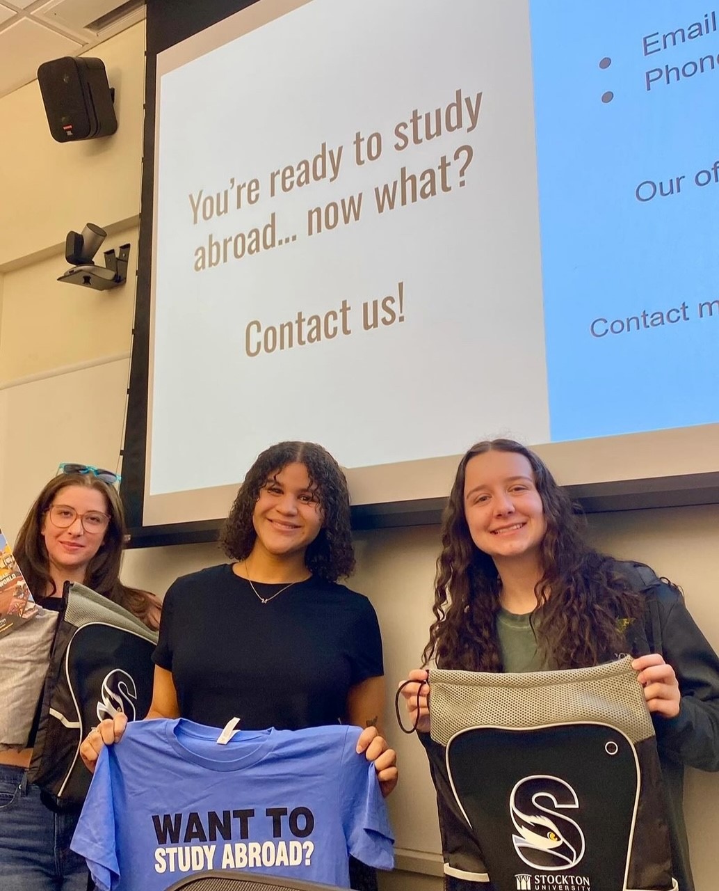 three students smiling, holding up study abroad shirts in front of a projector on the wall that states "You're ready to study abroad...now what?"
