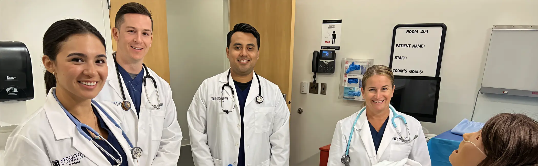 Four Stockton University nursing students in clinical coats and stethoscopes build advanced patient-care skills alongside a patient simulator in a nursing simulation room.