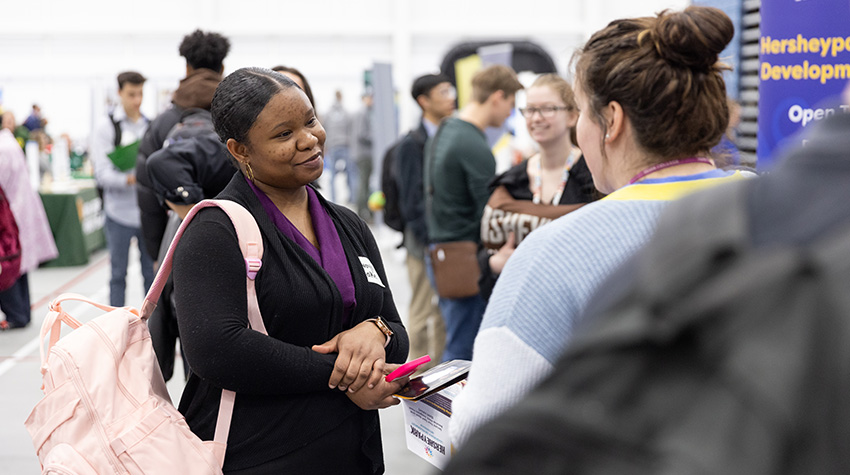 A student listening to a recruiter, holding a pink backpack on her arm. A student listening to a recruiter, holding a pink backpack on her arm.