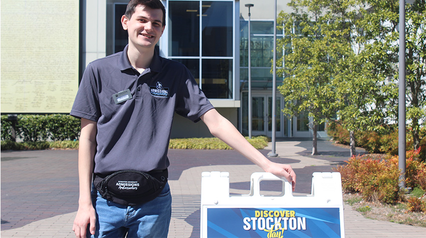 An Admissions Ambassador next to a sign that says Discover Stockton Day. An Admissions Ambassador next to a sign that says Discover Stockton Day.