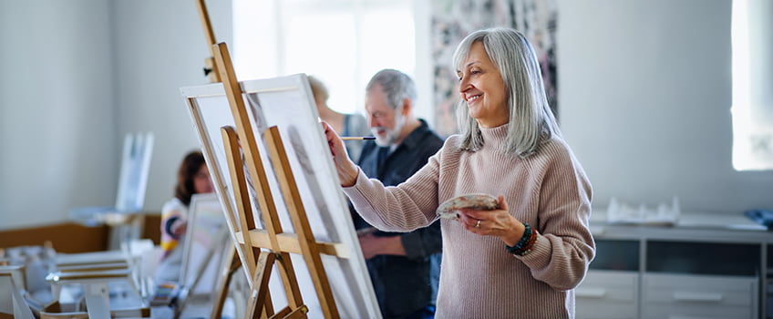 a woman is painting on a easel while others look on in the background and a man is holding a paintbrush a woman is painting on a easel while others look on in the background and a man is holding a paintbrush