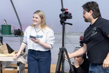 Team-based media shoot in a greenhouse setting, with students coordinating camera, audio, and setup.