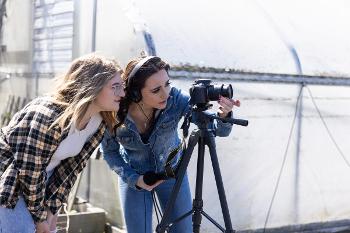 Two students film on location outside a greenhouse, planning shots and checking the DSLR on a tripod.