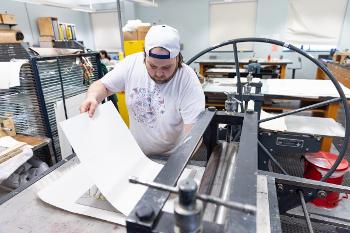 Printmaking student lifts a fresh print from the press, showcasing craftsmanship and hands-on studio learning.