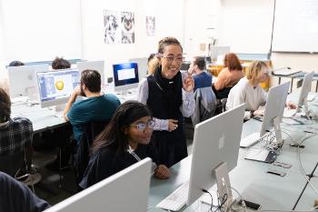 Faculty mentor guides students through digital illustration techniques in a computer lab using professional design tools.