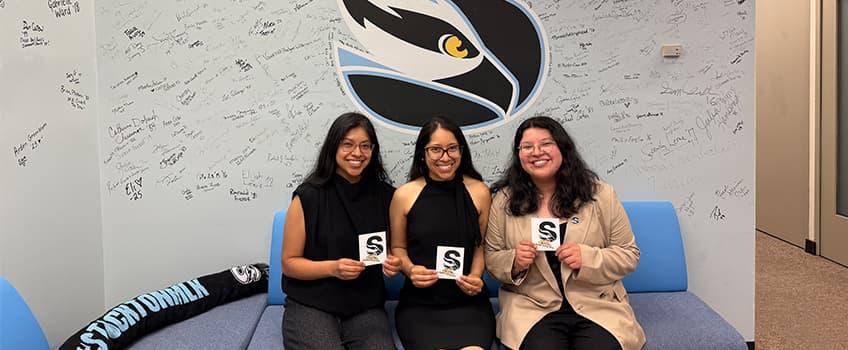 Three alumna holding Stockton "S" stickers and posing for a photo in front of the Alumni Hall of Fame Three alumna holding Stockton "S" stickers and posing for a photo in front of the Alumni Hall of Fame