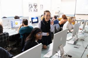 Faculty mentor guides students through digital illustration techniques in a computer lab using professional design tools. 