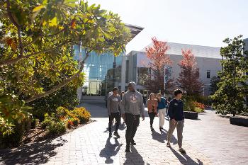 Stockton University students walk through a landscaped campus walkway surrounded by trees, gardens and modern academic buildings.