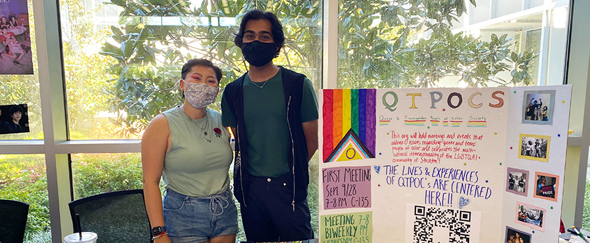 Mo Keane and Rikin Patel of QTPOCS at the Get Involved Fair; both are standing in behind a table and posterboard that says "QTPOCS: The lives and experiences of QTPOCSs are centered here!"