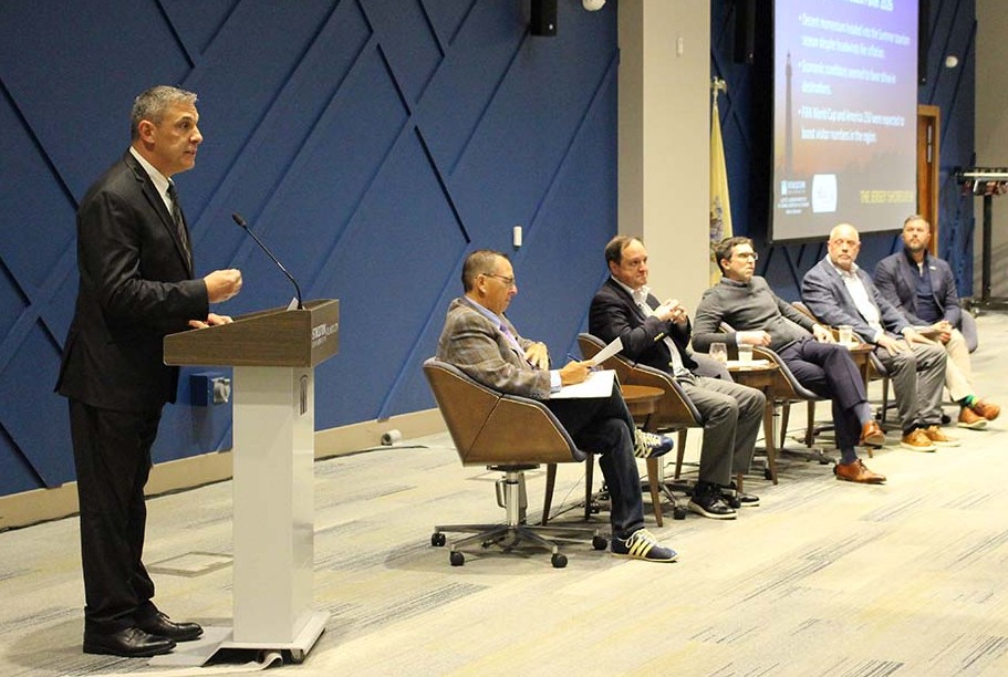 man speaks at podium next to seated panelists