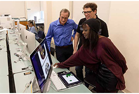 A student shows off her work on a computer screen to two faculty members