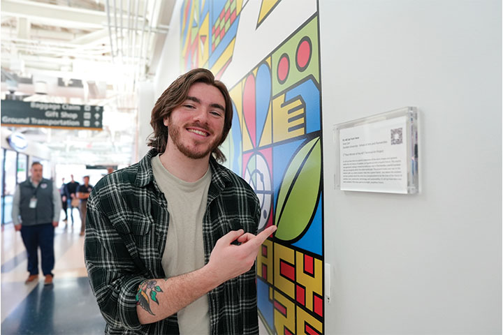 Evan Carr wears a green flannel shirt and points to his artwork hanging in an airport terminal