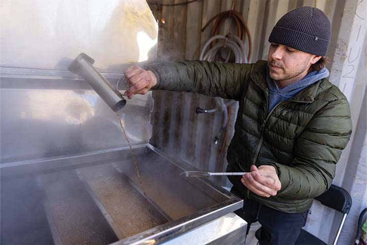 Ryan Hagerty in a green puffer jacket pours fresh maple syrup into a boiler.