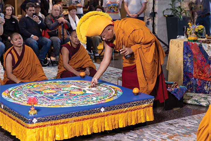 Three Tibetan monks in orange and red clothing stand around their colorful sand mandala