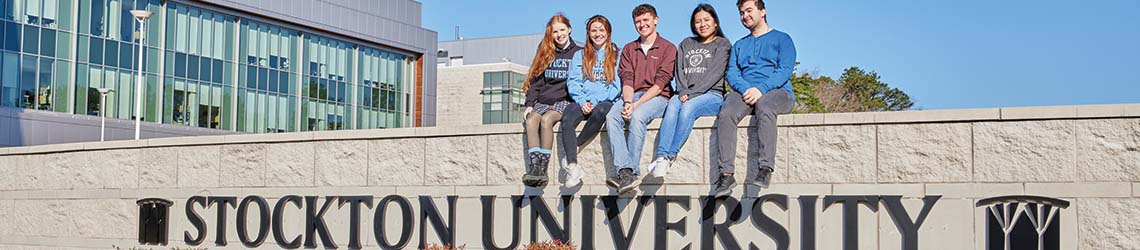 Five students sit on a white divider wall with Stockton University and the logo