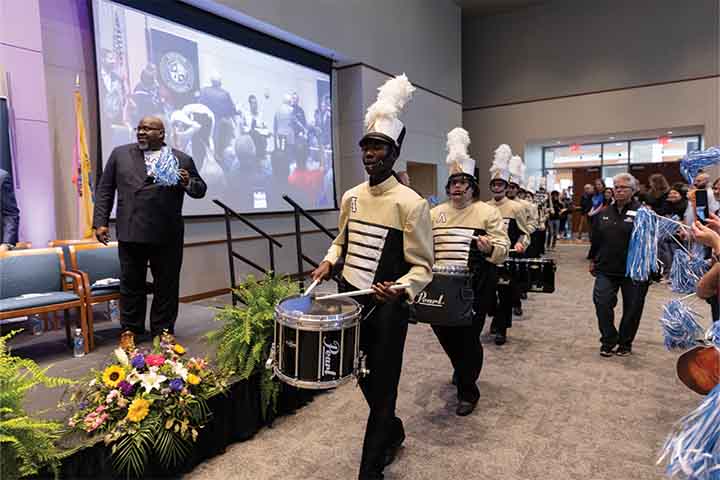 Drum line members wearing gold and black with white plume hats march into the Campus Center Event Room