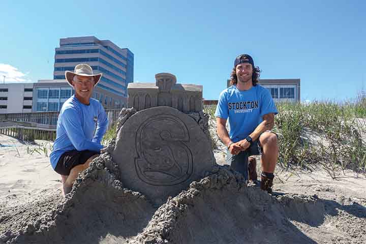 Matthew and Liam Deibert in blue Stockton shirts standing next to sand sculpture of the Campus Center and Stockton S