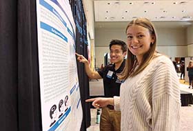 Two students point to their poster presentation hanging on a black curtain