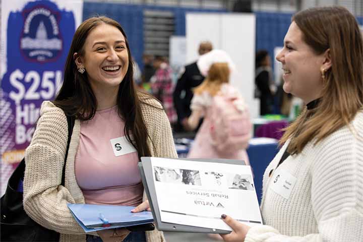 Two female students share a laugh while holding resumes and exploring employer tables at the career fair