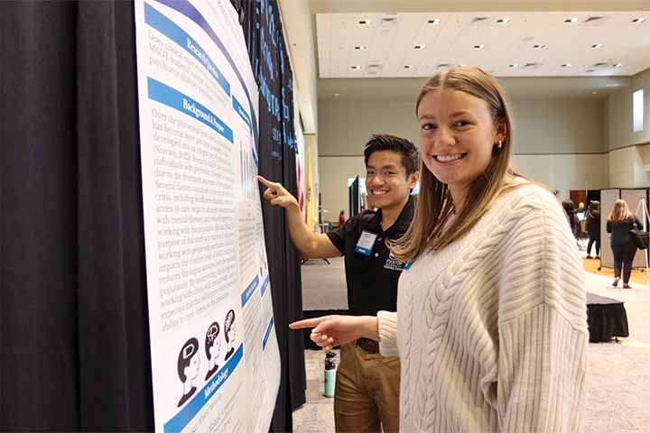 Two students point out information on their poster presentation hanging on a black curtain inside the Campus Center Event Room