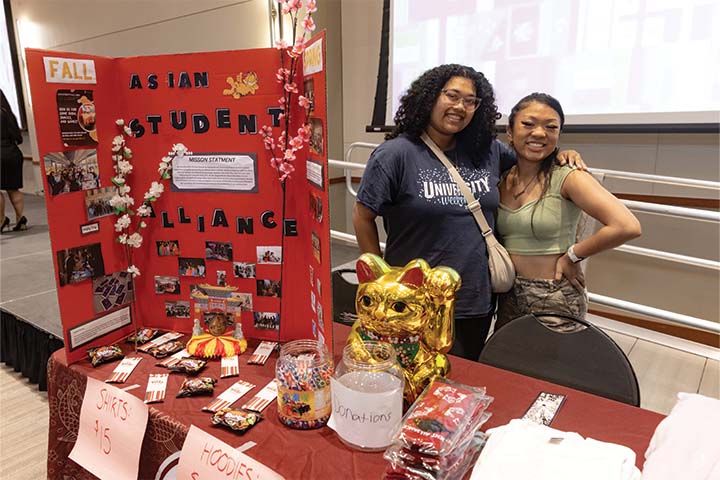 Two students pose and smile next to their table presentation about the Asian Student Alliance
